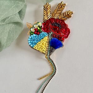 May include: A handcrafted brooch featuring a heart-shaped design with blue, yellow, and red beaded elements. The brooch includes gold-colored accents, a blue tassel, and dangling beaded strands, creating a unique and artistic accessory.