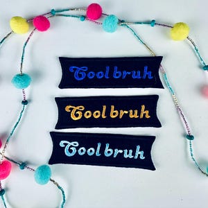 May include: Three black embroidered patches with the text "Cool bruh" in blue, gold, and light blue. A colorful garland with pom-poms and beads is in the background.