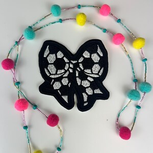 May include: A black bow-shaped patch with a soccer ball design, surrounded by a colorful beaded necklace. The necklace features pink, yellow, and teal felt balls, with small beads and turquoise accents.