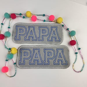 May include: Two gray felt patches with the word "PAPA" embroidered in blue. A beaded necklace with colorful pom-poms and beads in pink, yellow, and teal is also visible. The patches are rectangular with rounded corners.