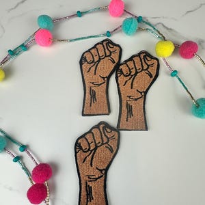 May include: Three brown fist patches with black outlines are displayed on a white surface. A beaded garland with pink, teal, and yellow pom-poms and beads is in the background. The patches are embroidered.