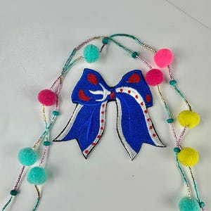 May include: A blue embroidered bow patch with white trim and red accents. The patch is surrounded by a beaded garland with colorful pom-poms in shades of pink, yellow, and teal. The design is set against a white background.