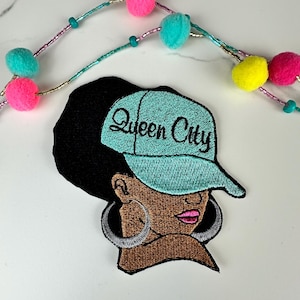 May include: Embroidered patch featuring a woman wearing a teal cap with the text "Queen City." The patch has a black background, brown skin, silver hoop earrings, and pink lipstick. A decorative beaded garland with pink, teal, and yellow pom-poms is in the background.