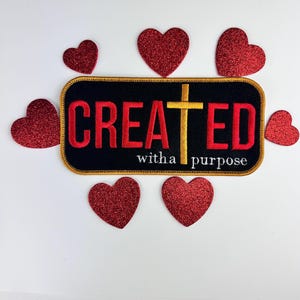 May include: A black and gold embroidered patch with the word "CREATED" in red letters and a gold cross. The text "with a purpose" is written in white below the word "CREATED". The patch is surrounded by red glitter hearts.
