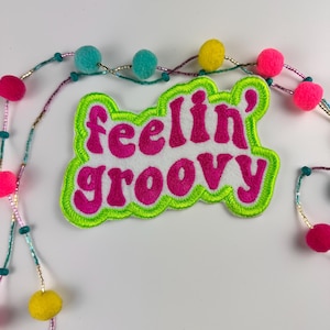 May include: A white patch with the words "feelin' groovy" in pink, outlined in neon green. The patch is surrounded by a beaded garland with pink, yellow, and teal pom-poms.