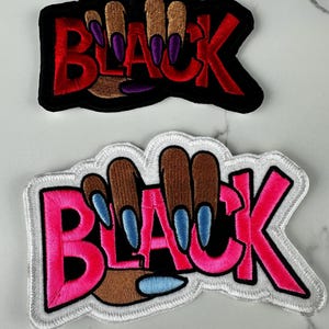 May include: Two embroidered patches featuring the word BLACK in bold lettering. The top patch has red letters with a black border and brown fingers with purple nails. The bottom patch has pink letters with a white border and brown fingers with blue nails.