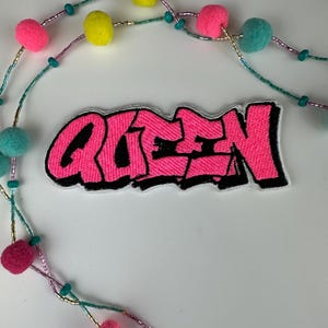 May include: A pink embroidered patch with the word "QUEEN" in a graffiti style, outlined in black. The patch is on a white surface, with a beaded necklace featuring pink, yellow, and teal pom-poms.