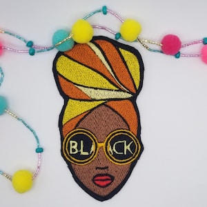 May include: Embroidered patch featuring a Black woman with a turban and sunglasses. The sunglasses have the word "BLACK" on them. The patch is surrounded by a beaded garland with yellow, pink, and teal pom-poms.
