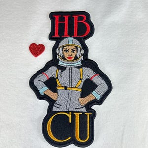 May include: Embroidered patch featuring an astronaut woman in a gray spacesuit with the letters "HB" above and "CU" below. A small red glitter heart is to the left of the patch. The astronaut has red lipstick and a yellow harness.