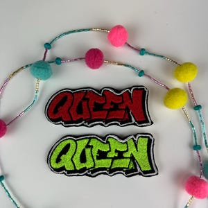 May include: Two embroidered patches with the word "QUEEN" in graffiti style, one red and one green. A beaded necklace with colorful pom-poms and beads in pink, yellow, and teal is also visible.