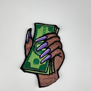 May include: Embroidered patch featuring a brown hand with long purple fingernails holding a stack of green $100 bills. The patch has a black border and is set against a white background. The design is a bold statement.