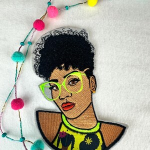 May include: Embroidered patch featuring a Black woman with curly black hair and neon green glasses. She wears a yellow top with a floral design. A colorful beaded necklace with pom-poms in pink, yellow, and teal accents the patch.