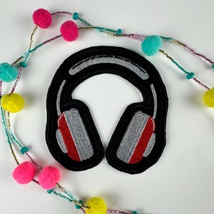 May include: Black embroidered patch in the shape of headphones with gray and red accents. The patch is surrounded by a colorful beaded garland with pink, yellow, and teal pom-poms.
