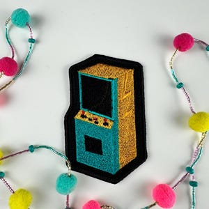 May include: Embroidered patch featuring a retro arcade game machine in teal, gold, and black. The patch has a black border and is surrounded by a colorful beaded garland with pom-poms in shades of pink, teal, and yellow.