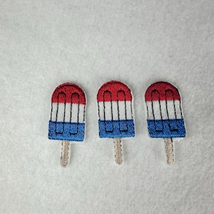 May include: Three embroidered popsicle patches. Each patch features a red top, white and black stripes, and a blue bottom. The popsicles have a light brown stick. The patches are on a white background.