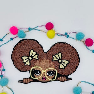 May include: Embroidered patch of a brown-skinned girl with large brown afro puffs, yellow bows, and yellow glasses peeking over a surface.
