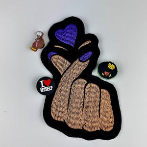 May include: A black patch with a brown hand making a heart shape with a purple heart on top. The patch is surrounded by three small colorful patches with the text "I love myself" on one of them.