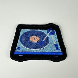 May include: Embroidered patch featuring a blue and light blue turntable design with a black border. The record player has a dark blue record with an orange center. The needle and other details are also blue.
