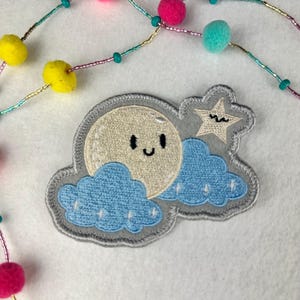 May include: An embroidered patch featuring a smiling moon, star, and blue clouds on a gray background. A beaded necklace with pink, yellow, and teal pom-poms and beads is in the background. The patch is a whimsical design.