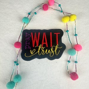 May include: A colorful necklace with pink, teal, and yellow pom-poms and beads. The necklace is paired with a black patch that reads "Pray Wait Trust" in red and gold lettering.