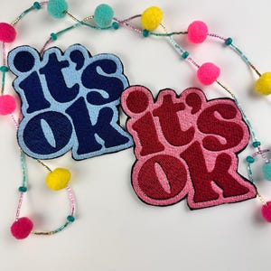 May include: Two embroidered patches with the words "it's ok" in a retro font. One patch is light blue with navy blue lettering, and the other is red with burgundy lettering. A beaded garland with colorful pom-poms and beads is in the background.