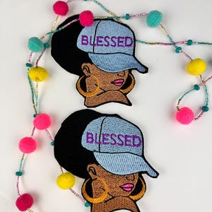 May include: Two embroidered patches featuring a Black woman wearing a light blue baseball cap with the word "BLESSED" in purple. The woman has large gold hoop earrings and pink lipstick. A beaded garland with colorful pom-poms and beads is in the background.