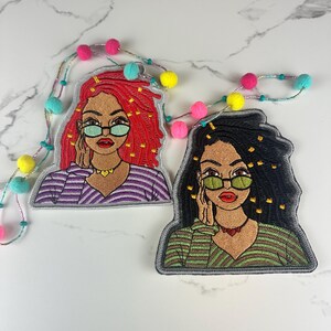 May include: Two embroidered patches featuring stylized portraits of women with glasses. One has red hair and a purple striped shirt, the other has black hair and a green striped shirt. Both have a gold necklace and are framed with a gray border.
