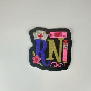 May include: Embroidered patch featuring the letters "RN" in gold and blue, with a pink syringe, a nurse's cap with a red cross, and a pink bandage. A pink flower and decorative swirls are also included.