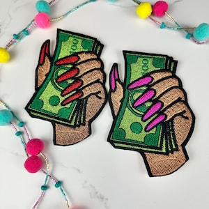 May include: Two embroidered patches featuring hands holding stacks of green $100 bills. One patch has red fingernails, the other has pink fingernails. The patches are outlined in black and have a brown skin tone.