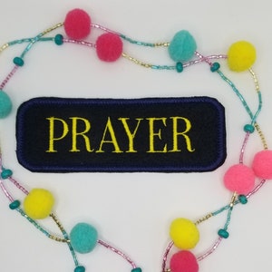 May include: A navy blue embroidered patch with the word "PRAYER" in yellow, surrounded by a beaded garland with colorful pom-poms. The garland features pink, yellow, and teal pom-poms, and teal and gold beads.