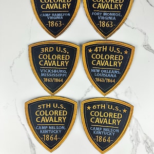 May include: Six shield-shaped embroidered patches with black fabric and gold borders. Each patch features gold text identifying units from the 1st through 6th U.S. Colored Cavalry, including locations like Camp Hamilton, Fort Monroe, Vicksburg, New Orleans, and Camp Nelson, with dates ranging from 1863 to 1864.