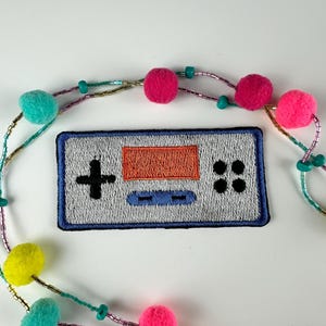 May include: Embroidered patch featuring a retro video game controller design with a gray background, orange screen, and blue accents. A colorful beaded necklace with pink, teal, and yellow pom-poms and beads surrounds the patch.