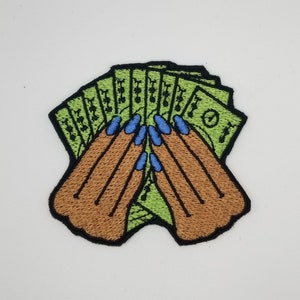 May include: Embroidered patch featuring a fan of green money with brown hands and blue fingernails. The patch has a black border and is set against a white background. The design is a stylized representation of wealth.