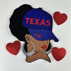 May include: Embroidered patch featuring a Black woman wearing a blue baseball cap with "TEXAS" in red lettering. The patch includes the number "254" and is surrounded by three red glitter hearts. The woman has large silver hoop earrings.
