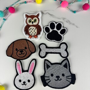 May include: Embroidered patches featuring an owl, dog, paw print, bone, bunny, and cat. The patches are in various colors, including brown, black, and white, with detailed stitching. A colorful beaded garland with pom-poms is in the background.