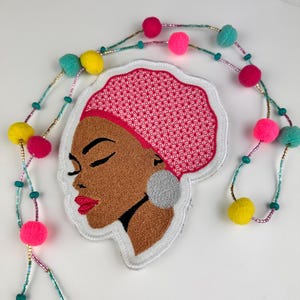 May include: Embroidered patch featuring a woman's profile with closed eyes, red lips, and a pink head wrap. A beaded garland with colorful pom-poms and beads surrounds the patch. The woman has a silver earring.