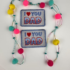 May include: Two embroidered patches with the text "I ❤️ YOU DAD" in blue and red, and two beaded necklaces with colorful pom-poms. The pom-poms are yellow, pink, and teal. The necklaces have small beads and are on a white background.