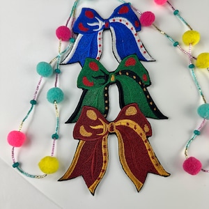 May include: Three embroidered bow patches in blue, green, and burgundy, each with a unique design. A beaded garland with colorful pom-poms and beads is draped across the image. The bows are likely for crafts or embellishments.