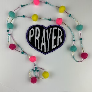 May include: A purple heart-shaped patch with the word "PRAYER" embroidered in white on a black background. The patch is attached to a beaded necklace with pink, yellow, and turquoise pom-poms.