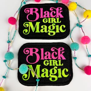 May include: Two black iron-on patches with the text "Black Girl Magic" in pink and green embroidery. The patches are on a white background with a colorful beaded garland.
