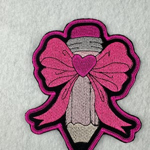 May include: Embroidered patch featuring a pencil design with a pink bow and heart. The pencil is light gray with a pink eraser. The bow and heart are bright pink with a black outline. The patch has a black border.