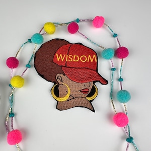 May include: An embroidered patch featuring a woman wearing a red cap with the word "WISDOM" in gold lettering. The woman has brown skin, gold hoop earrings, and brown hair. A beaded garland with pink, yellow, and turquoise pom-poms and beads surrounds the patch.