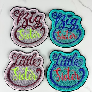 May include: Four embroidered patches with black borders. Two patches are light pink with dark maroon text reading Big Sister and Little Sister in lime green. Two patches are teal with dark blue text reading Big Sister and Little Sister in red.