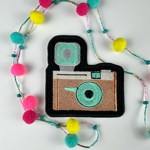 May include: A colorful necklace with a camera patch. The patch is brown, black, and mint green. The necklace has a beaded strand with pink, yellow, and teal pom-poms.