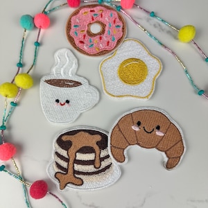 May include: Embroidered patches of breakfast foods, including a pink donut with sprinkles, a fried egg, a coffee cup, pancakes with syrup, and a croissant. The patches are colorful and have smiling faces.