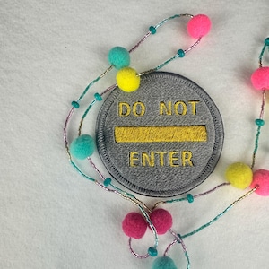 May include: A gray embroidered patch with the words "DO NOT ENTER" in yellow. The patch is surrounded by a beaded string with colorful pom-poms in pink, teal, and yellow.
