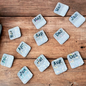 May include: Twelve small, white, square cards with the months of the year printed on them. The cards are scattered on a wooden surface.