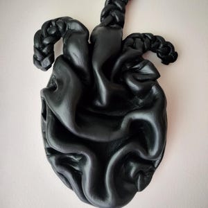 May include: A black, sculpted heart-shaped wall hanging. The heart has a textured, organic appearance with braided elements at the top. The artwork is mounted on a light pink wall, creating a contrast with the dark sculpture.