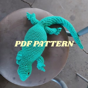 May include: A crocheted green lizard toy with a long tail. The toy is sitting on a brown stool. The text "PDF PATTERN" is visible on the stool.
