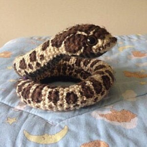 May include: A crocheted snake toy in shades of brown and cream. The snake is coiled, displaying a detailed spot pattern and a small, dark eye. It rests on a blue patterned surface.